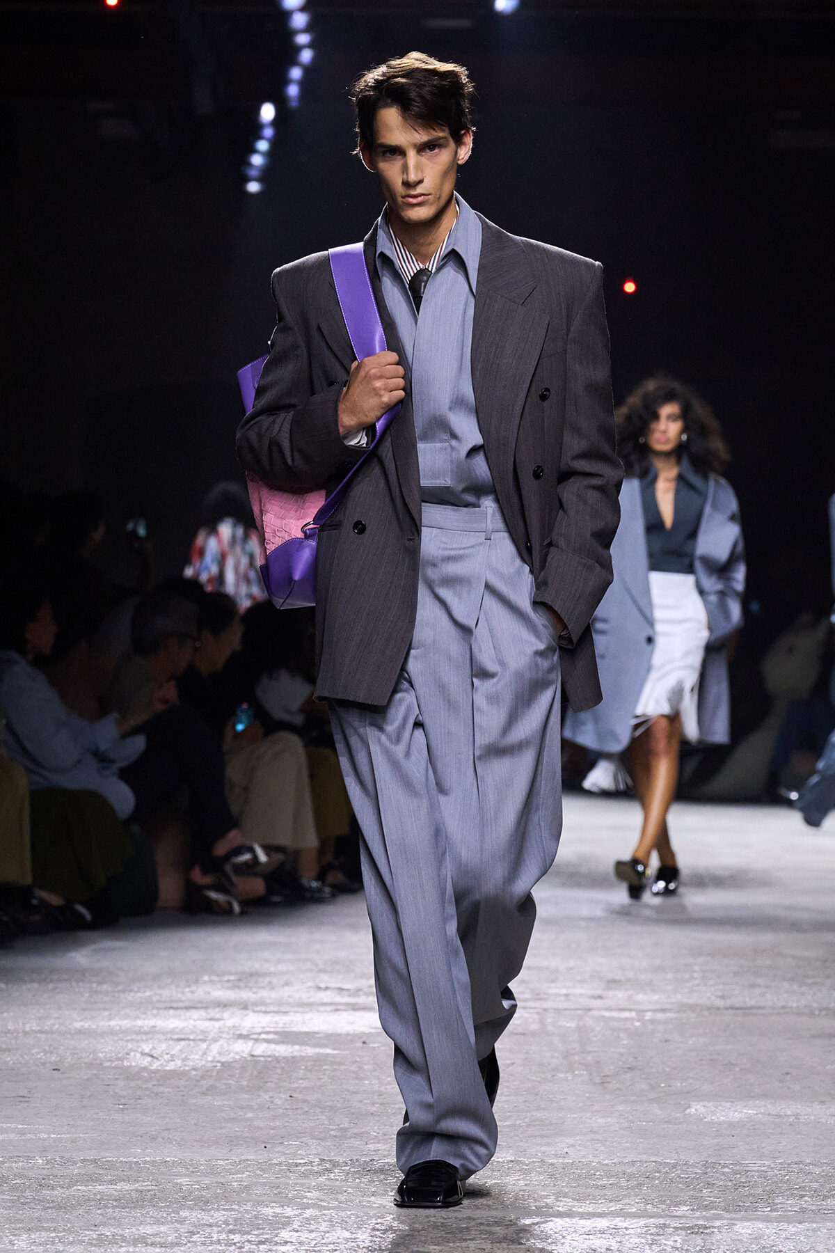 Male model walks the runway in an oversized gray suit with a light blue shirt, striped tie, and a purple shoulder bag.