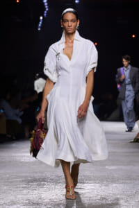 Runway model in a white midi dress with rolled sleeves, walking forward with a colorful woven handbag in hand.