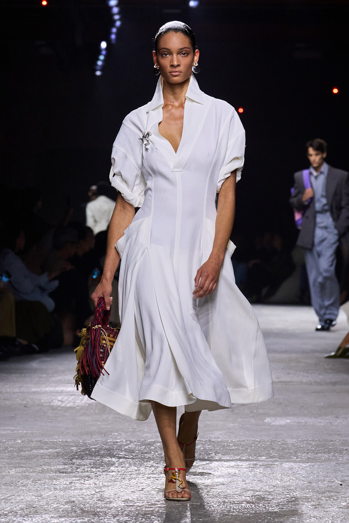 Runway model in a white midi dress with rolled sleeves, walking forward with a colorful woven handbag in hand.