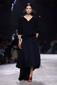 Model walks the runway in a black knit sweater with a V-neck and a navy pleated midi skirt, paired with red open-toe sandals.
