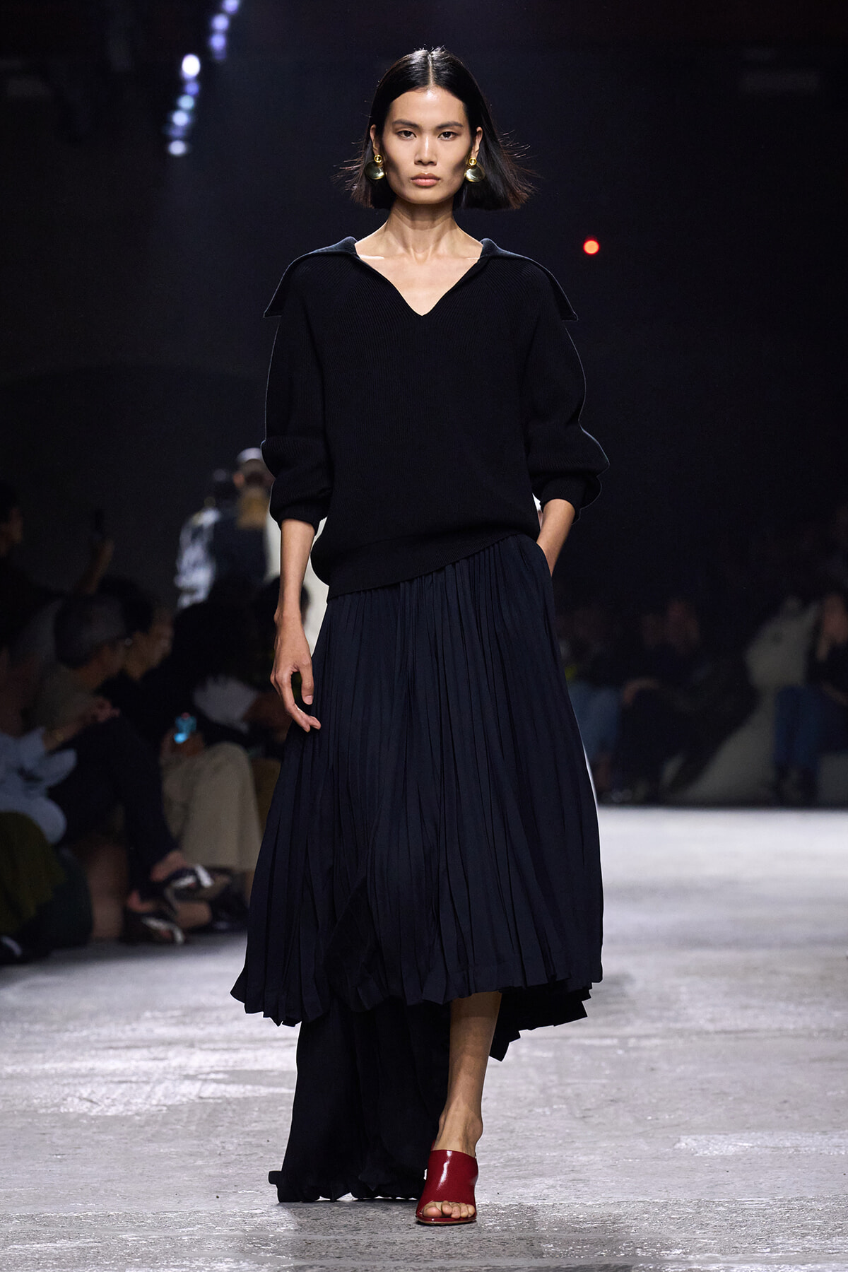 Model walks the runway in a black knit sweater with a V-neck and a navy pleated midi skirt, paired with red open-toe sandals.