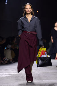 Model walks the runway in a gray blouse and burgundy asymmetric wrap skirt with a belt, holding a yellow bag with a rabbit graphic and a black handbag.