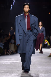 Runway model in an oversized blue suit with a burgundy shirt and pale tie, walking the catwalk with an audience in the background.