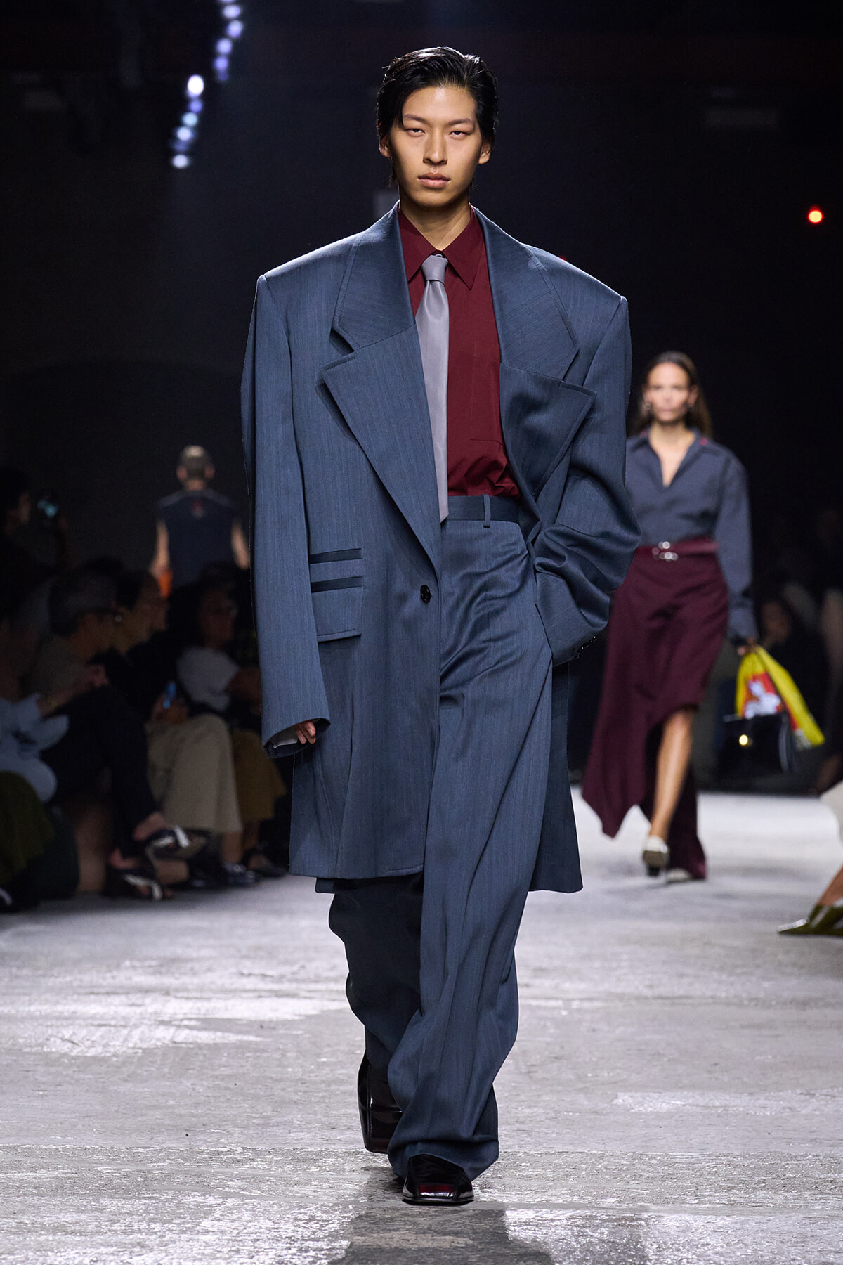 Runway model in an oversized blue suit with a burgundy shirt and pale tie, walking the catwalk with an audience in the background.