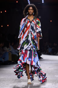 Model walking a fashion runway in a bold, multicolor abstract-print dress with cascading ruffles and white open-toe heels.