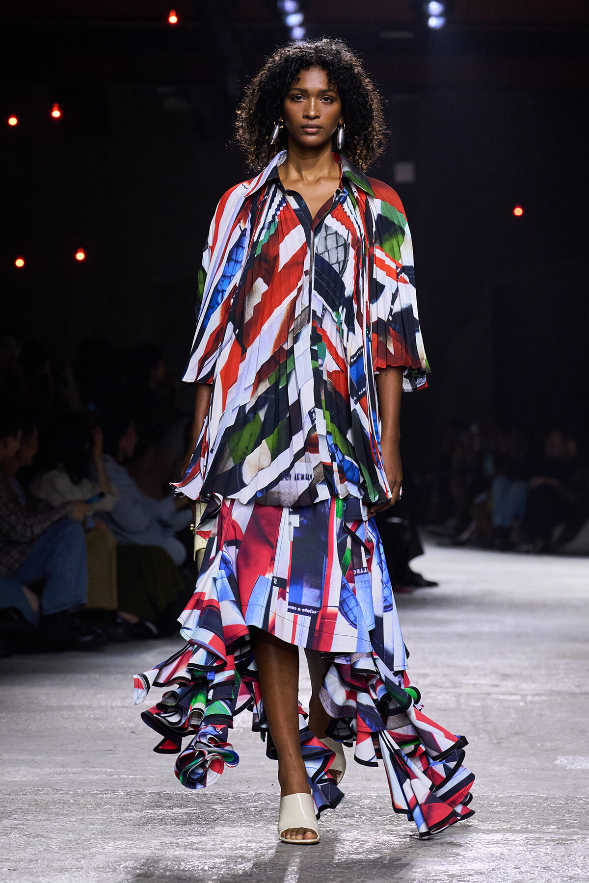 Model walking a fashion runway in a bold, multicolor abstract-print dress with cascading ruffles and white open-toe heels.