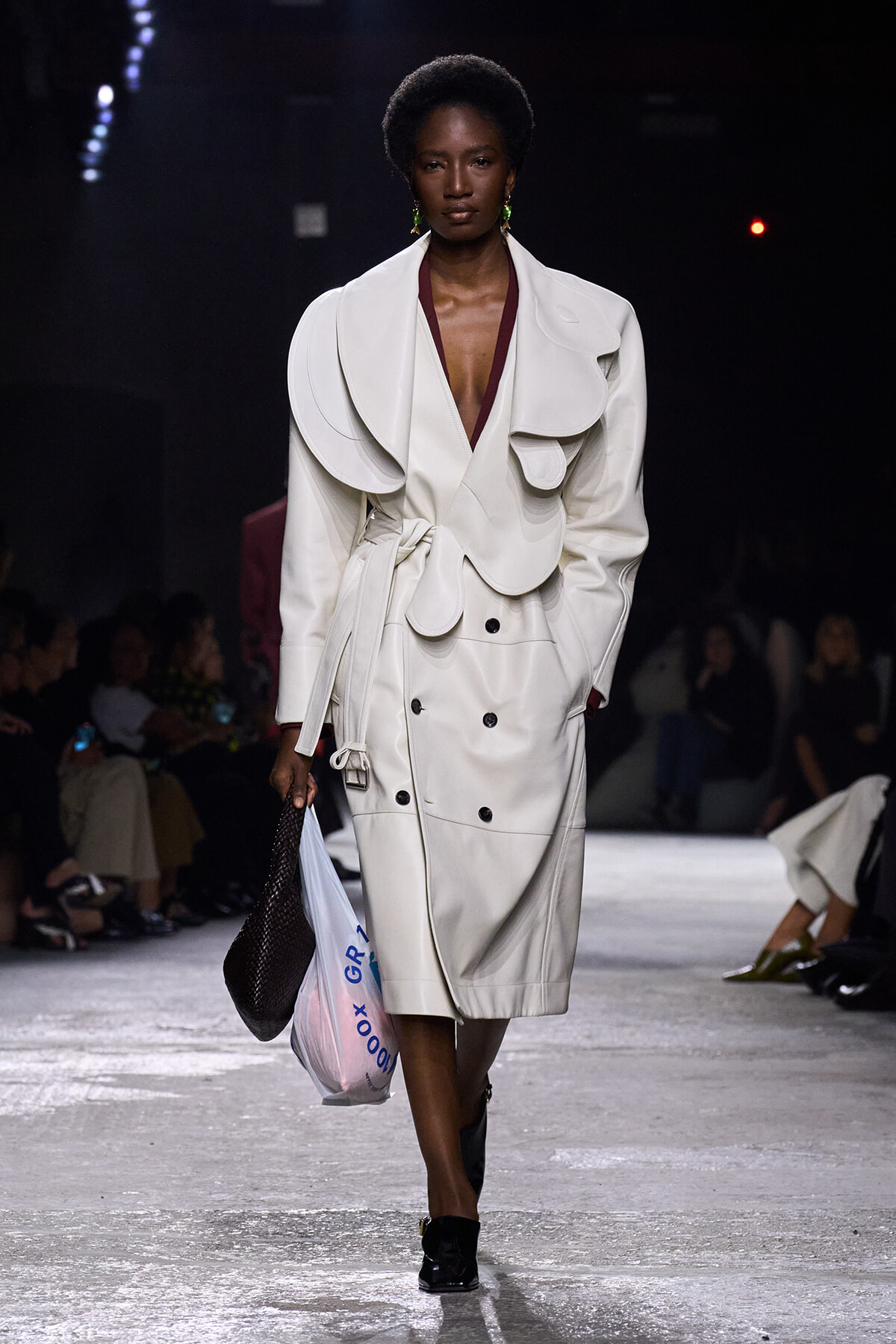Model walks the runway in a white structured trench with oversized sculptural shoulders and a deep V-neck, belt tied at the waist, carrying a brown woven bag and a plastic shopping bag.