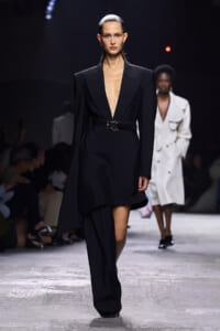 Model walks the runway in a black deep-plunge blazer dress with wide shoulders and a waist belt.