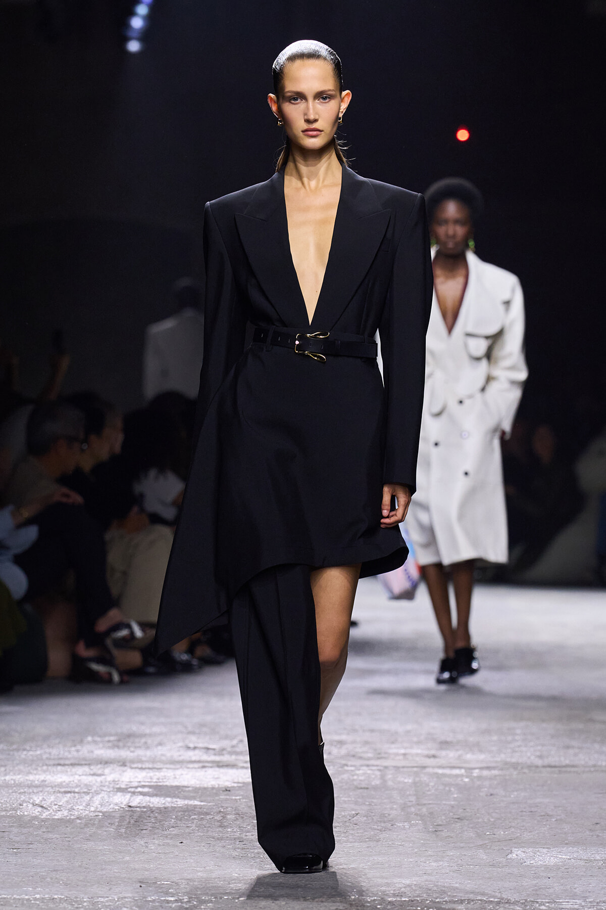 Model walks the runway in a black deep-plunge blazer dress with wide shoulders and a waist belt.