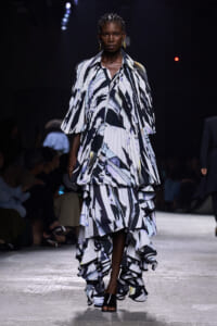 Female model walks a runway in a voluminous black-and-white abstract print gown with layered ruffles and cape-like sleeves, under dark stage lighting.