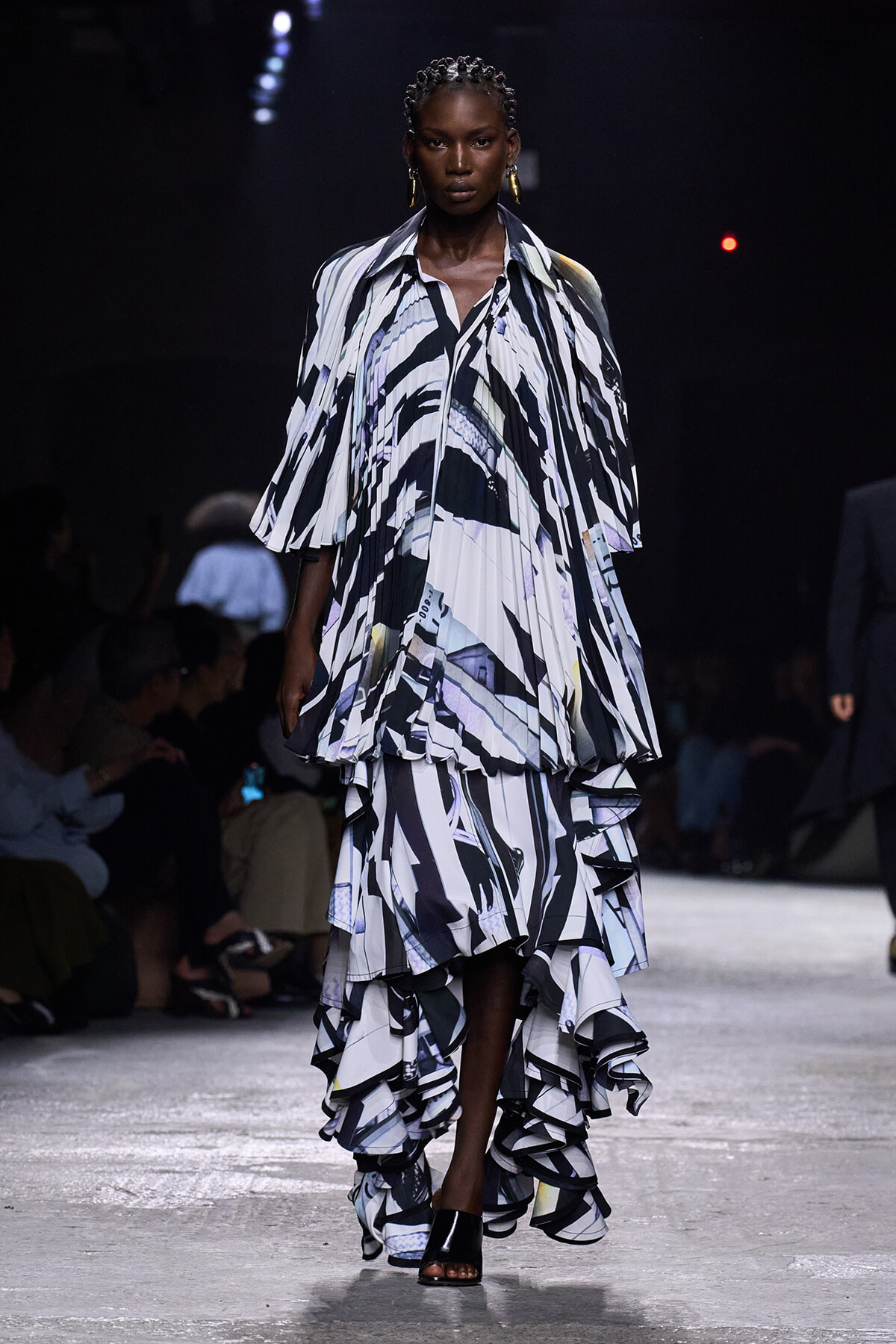 Female model walks a runway in a voluminous black-and-white abstract print gown with layered ruffles and cape-like sleeves, under dark stage lighting.