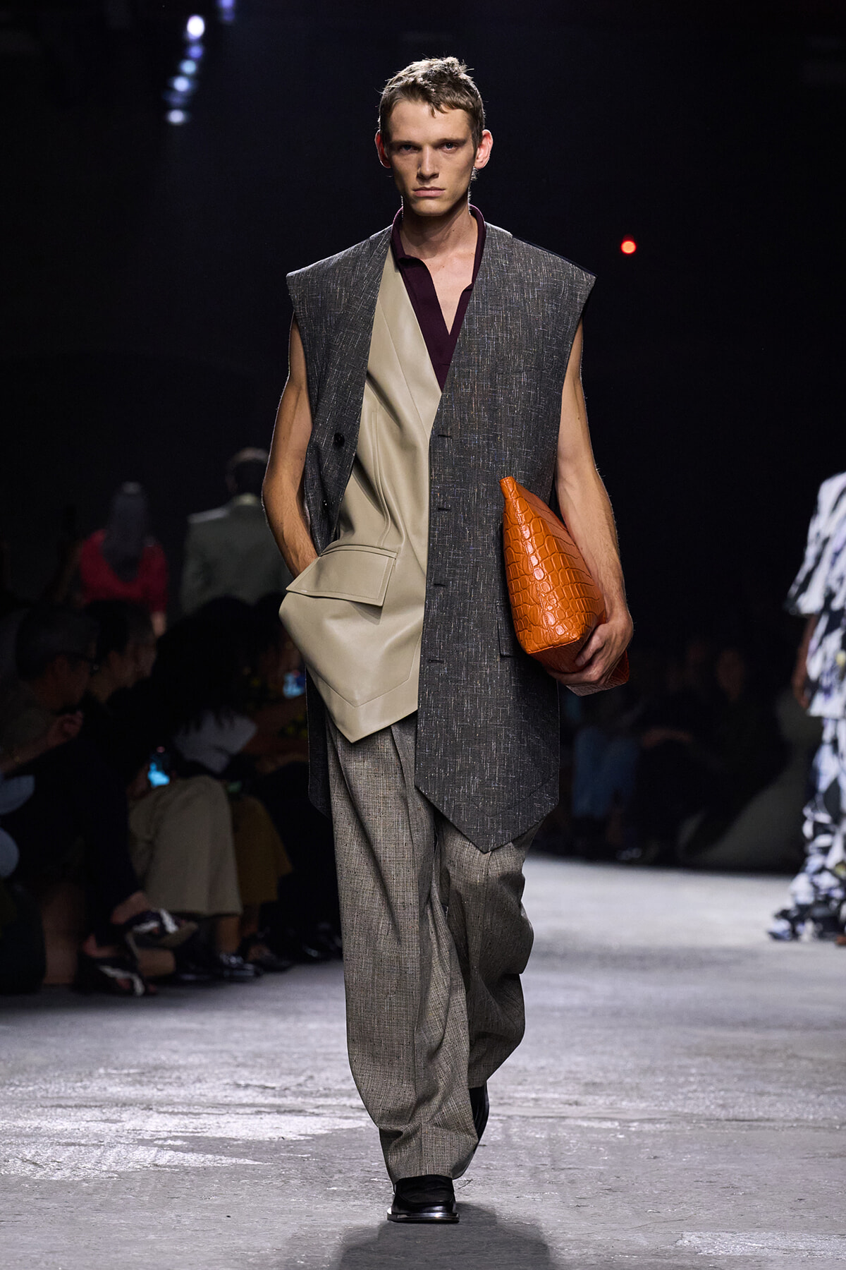 Male model walks the runway in a layered gray vest over beige blazer and loose gray trousers, carrying an orange textured clutchbag.