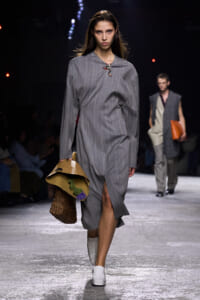 Model walking a runway in a gray pinstriped midi dress with a front slit, carrying a tan hat and a wicker bag.