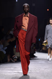 Runway model wears a burgundy oversized blazer over an orange shirt and red wide-leg pants, carrying a dark clutch.