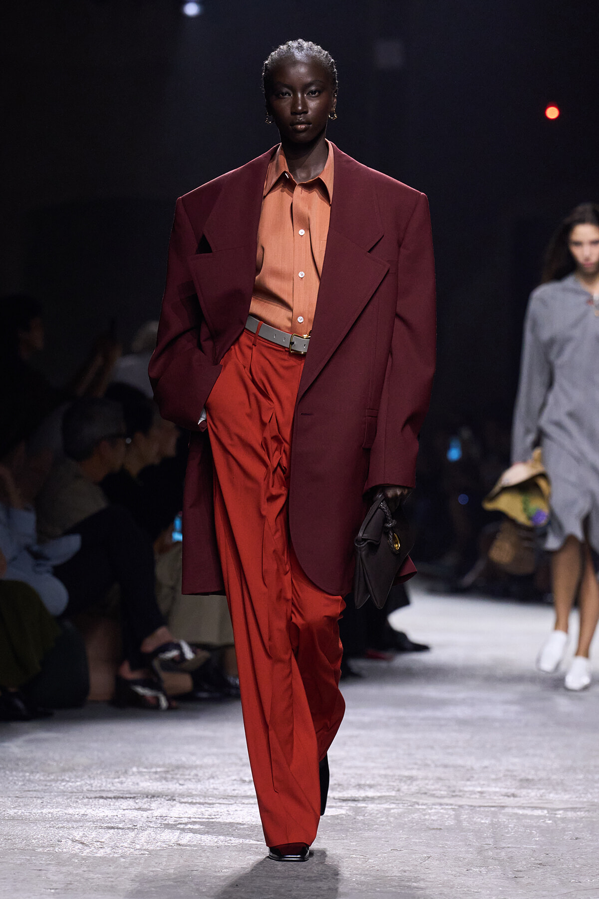 Runway model wears a burgundy oversized blazer over an orange shirt and red wide-leg pants, carrying a dark clutch.