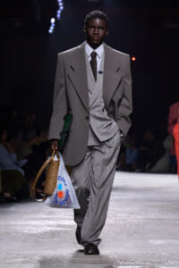 Male model walking a runway in an oversized gray suit with a layered vest, striped shirt, and dark tie, carrying a woven basket bag and a white plastic shopping bag.