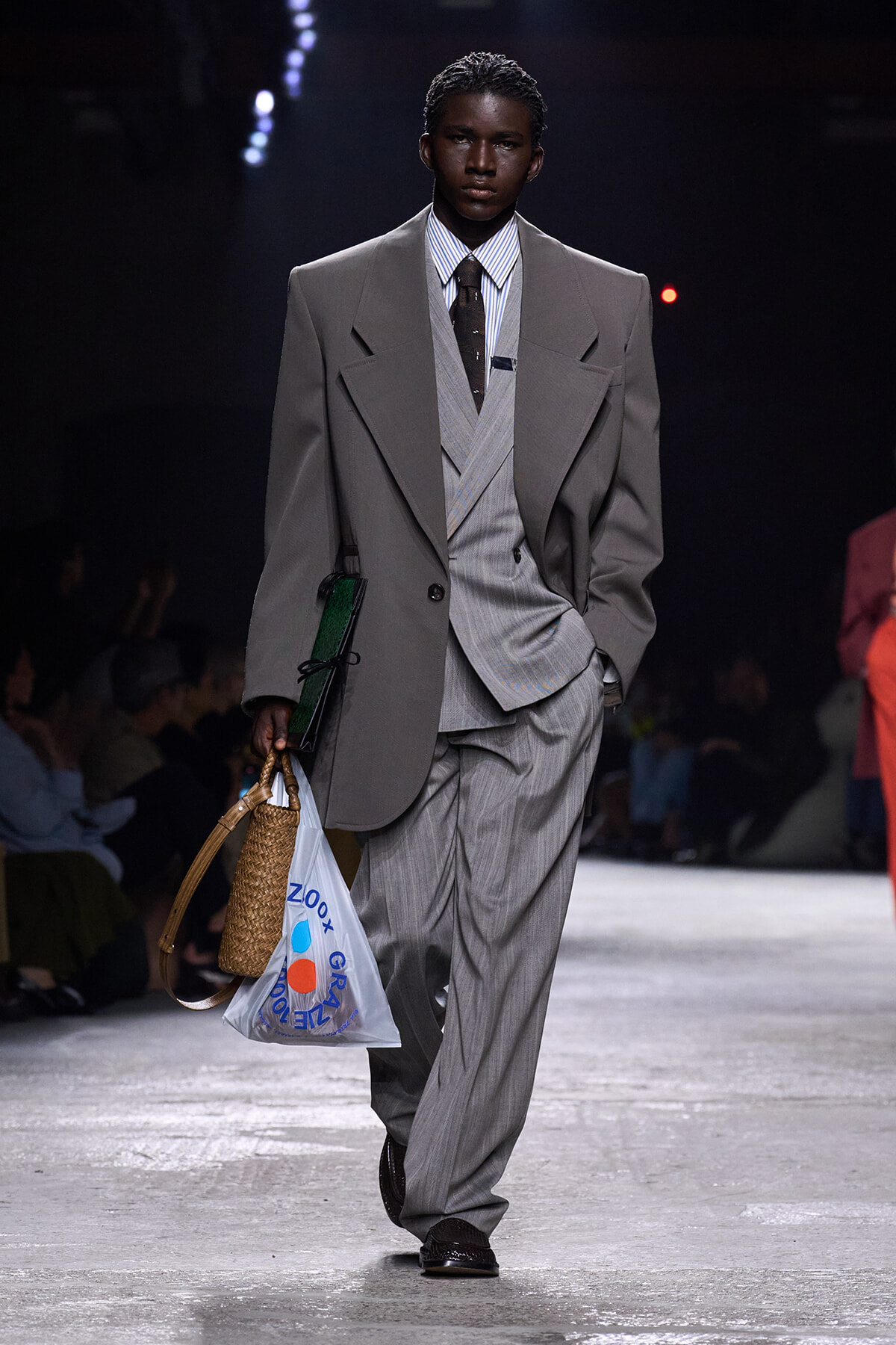 Male model walking a runway in an oversized gray suit with a layered vest, striped shirt, and dark tie, carrying a woven basket bag and a white plastic shopping bag.