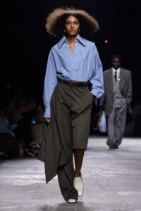 Model walking runway in a blue striped oversized shirt tucked into a dark olive asymmetrical wrap skirt with a belt and white ankle boots.