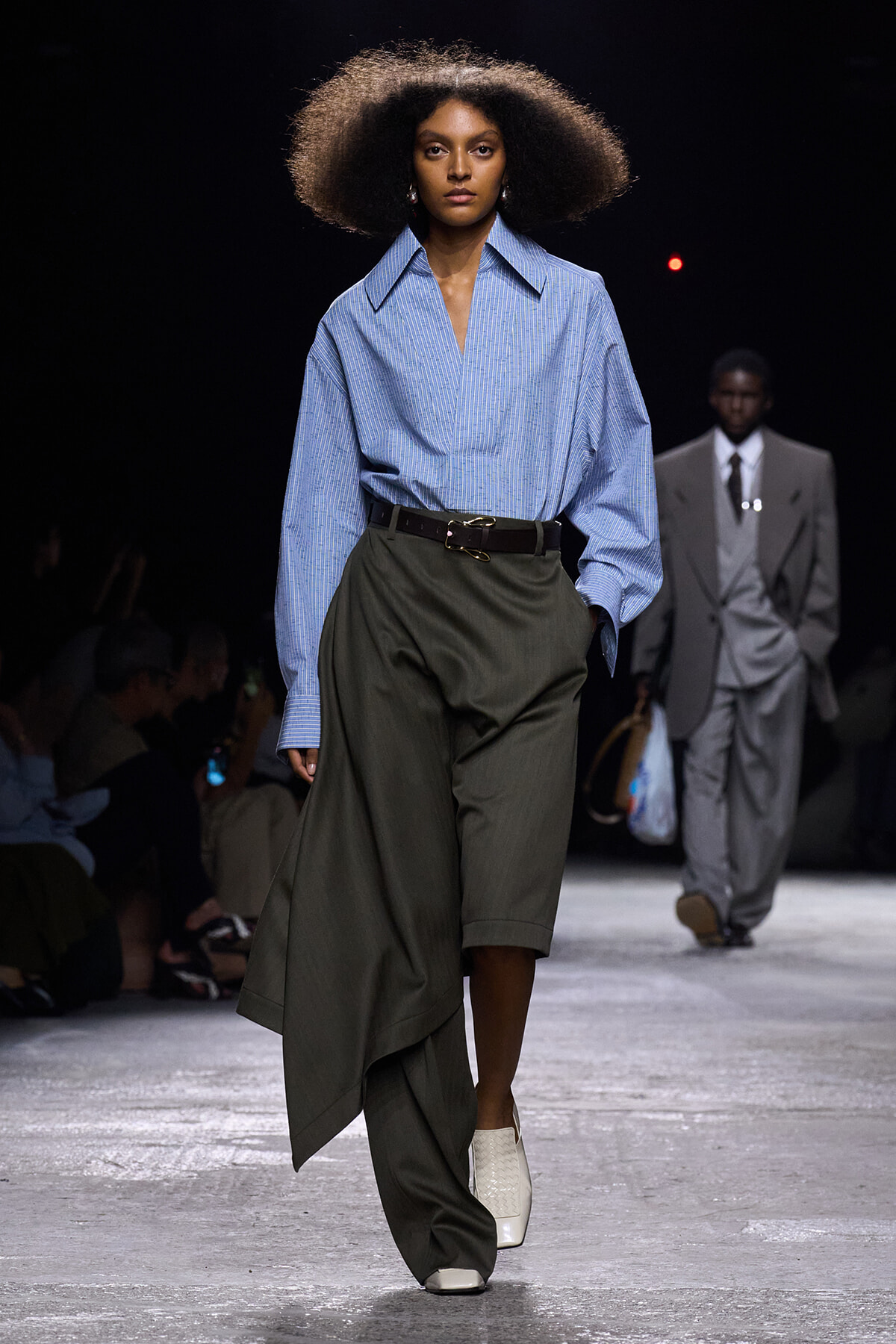 Model walking runway in a blue striped oversized shirt tucked into a dark olive asymmetrical wrap skirt with a belt and white ankle boots.