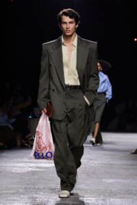 Male runway model wearing a loose olive suit with a cream shirt, belt, and white shoes, carrying a pink plastic shopping bag.