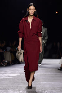 Model walks the runway in a burgundy belted jumpsuit with rolled sleeves, carrying a beige crocodile-textured handbag and wearing dark heels.