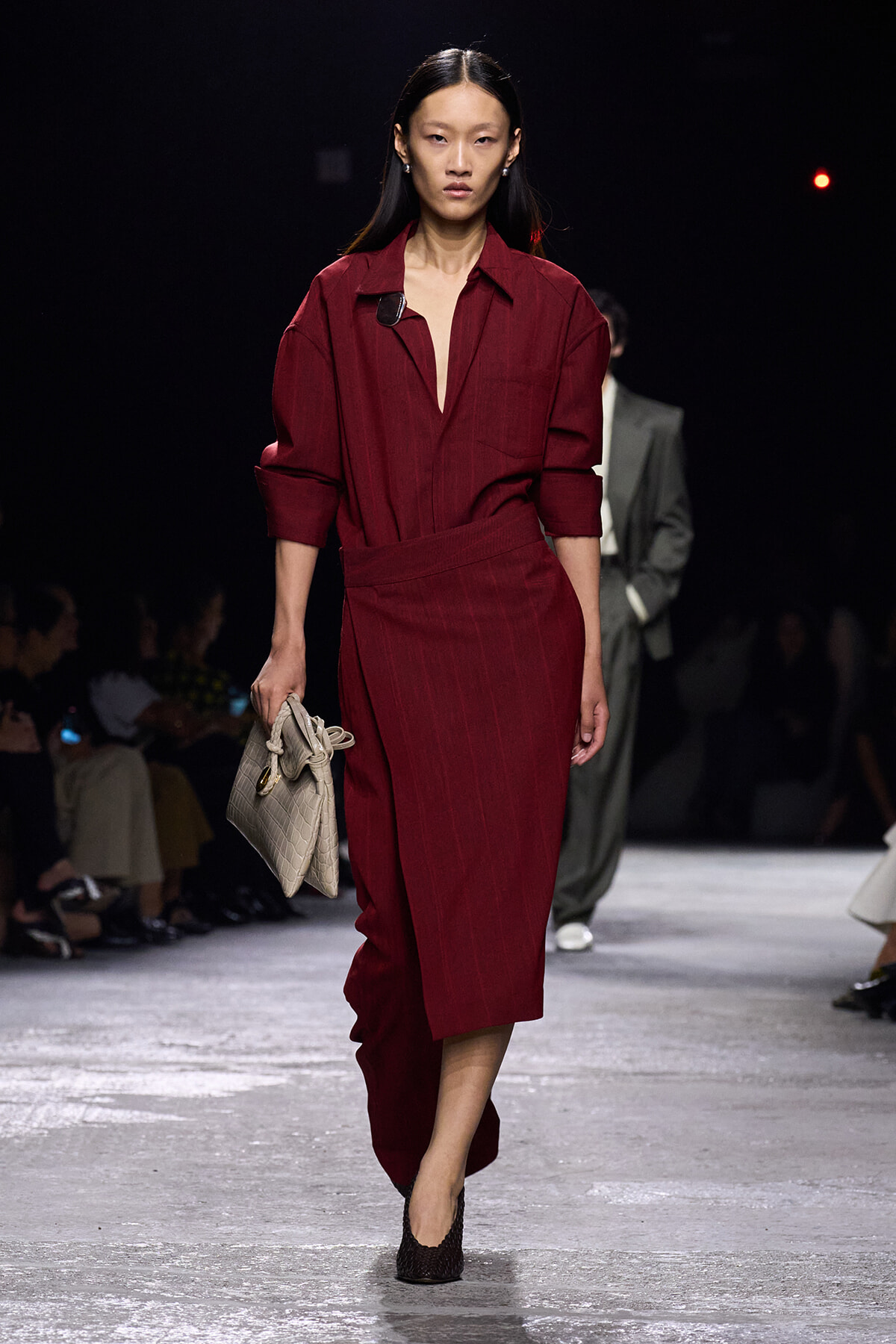 Model walks the runway in a burgundy belted jumpsuit with rolled sleeves, carrying a beige crocodile-textured handbag and wearing dark heels.