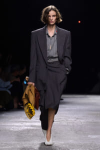 Runway model in a charcoal pinstripe suit with a grey blouse, belt, and white textured shoes, carrying a tan coat with a sunflower accessory.