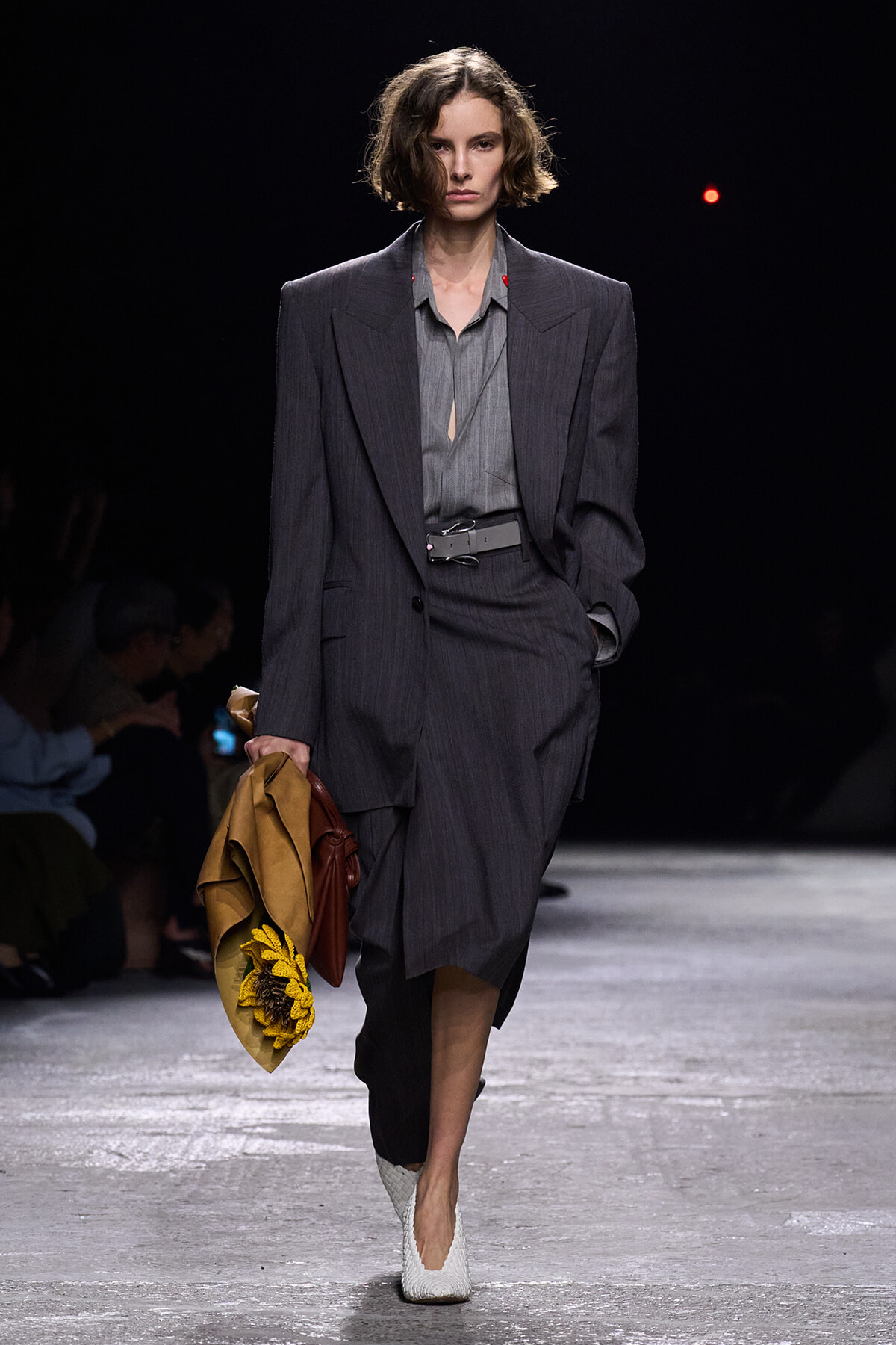 Runway model in a charcoal pinstripe suit with a grey blouse, belt, and white textured shoes, carrying a tan coat with a sunflower accessory.