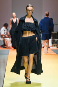 Model walks the runway in an all-black ensemble: a black knitted long coat over a ruffled crop top and matching shorts, with sandals.