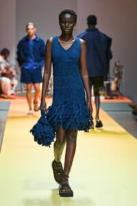 Model walking a fashion show in a blue textured dress with a tiered fringe hem, carrying a matching fringed bag.