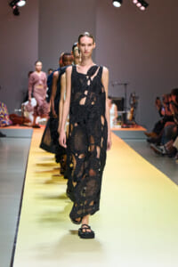 Model walks a yellow runway in a black, cut-out, mesh dress with circular patterns and platform sandals.