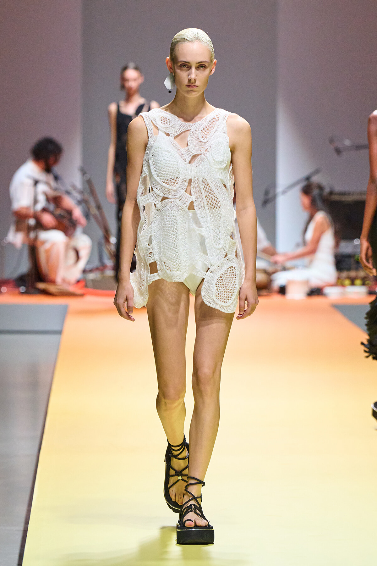 Model walks the runway in a white crocheted sleeveless dress with intricate cutouts and black lace-up sandals.
