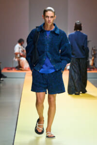 Male model walking a runway in a textured blue knit cardigan and matching shorts, wearing white sandals and carrying a dark bag on his shoulder.