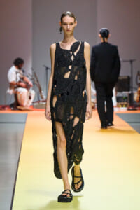 Model walking a fashion show runway in a black cut‑out mesh dress with irregular holes, wearing sandals.