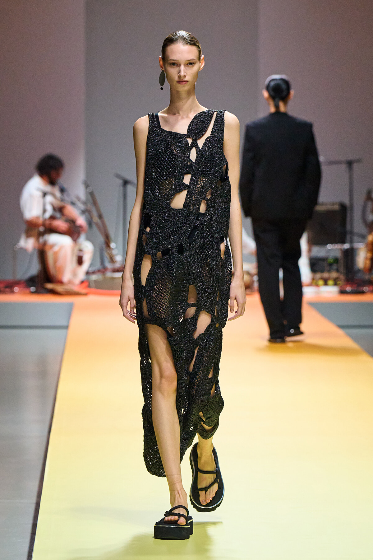 Model walking a fashion show runway in a black cut‑out mesh dress with irregular holes, wearing sandals.
