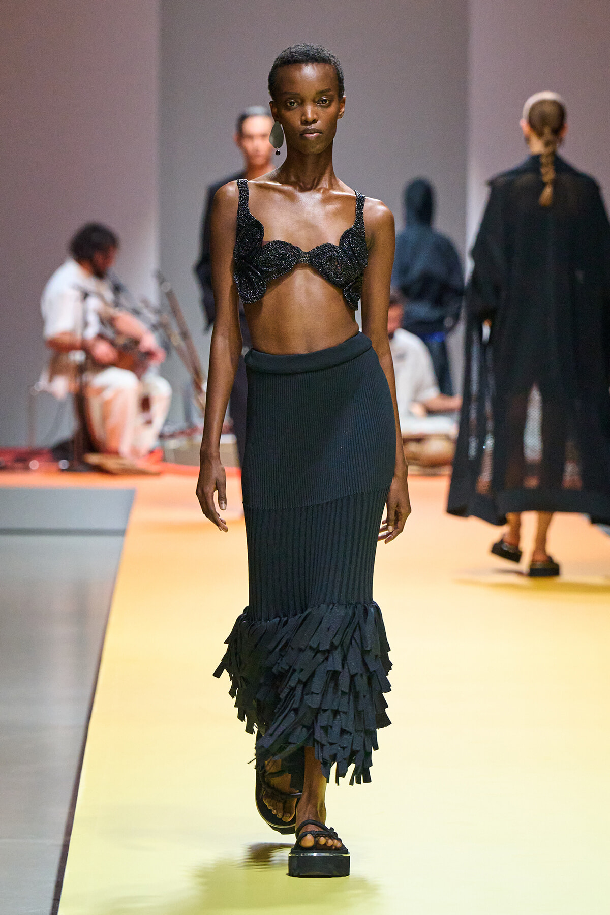 Model walks the runway in a black beaded bralette top and a long ribbed black skirt with a layered fringe hem, on a yellow catwalk.