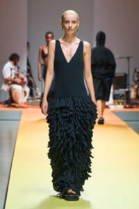 Model walks the runway in a sleeveless black V-neck dress with a floor-length fringe skirt made of layered strips of fabric.