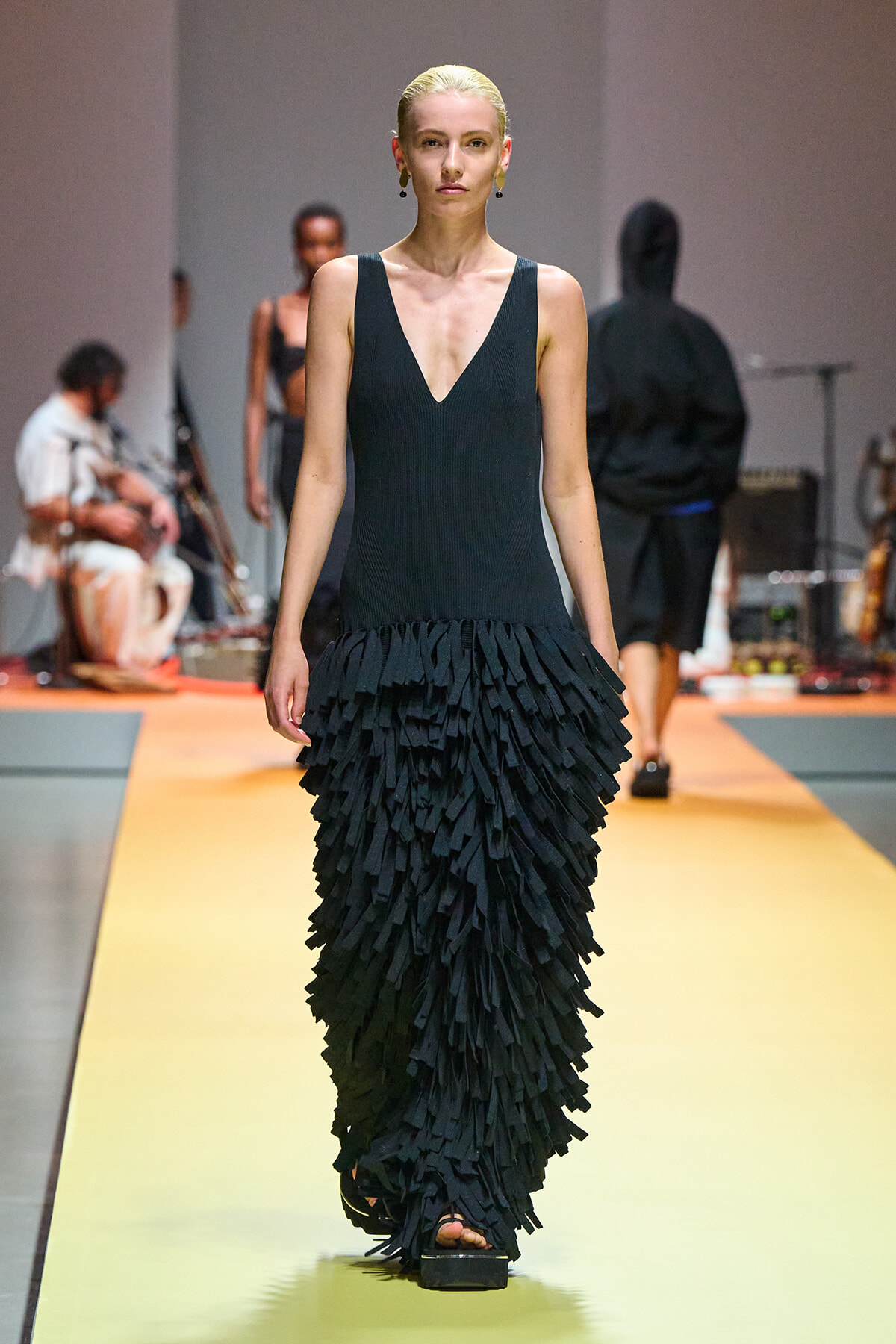 Model walks the runway in a sleeveless black V-neck dress with a floor-length fringe skirt made of layered strips of fabric.