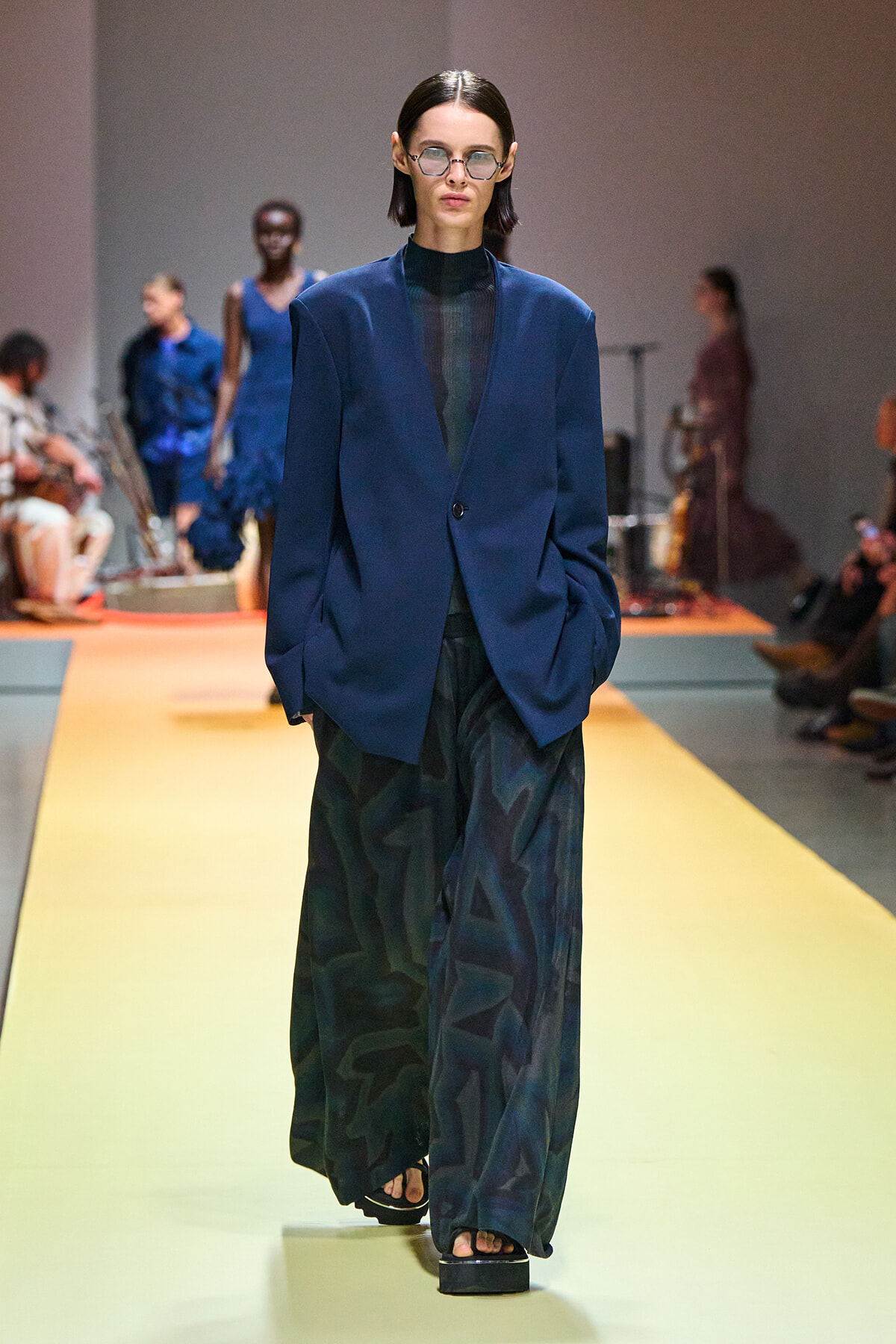 Model walks the runway in a blue blazer, dark sheer top, and wide, dark patterned pants with platform sandals and sunglasses.