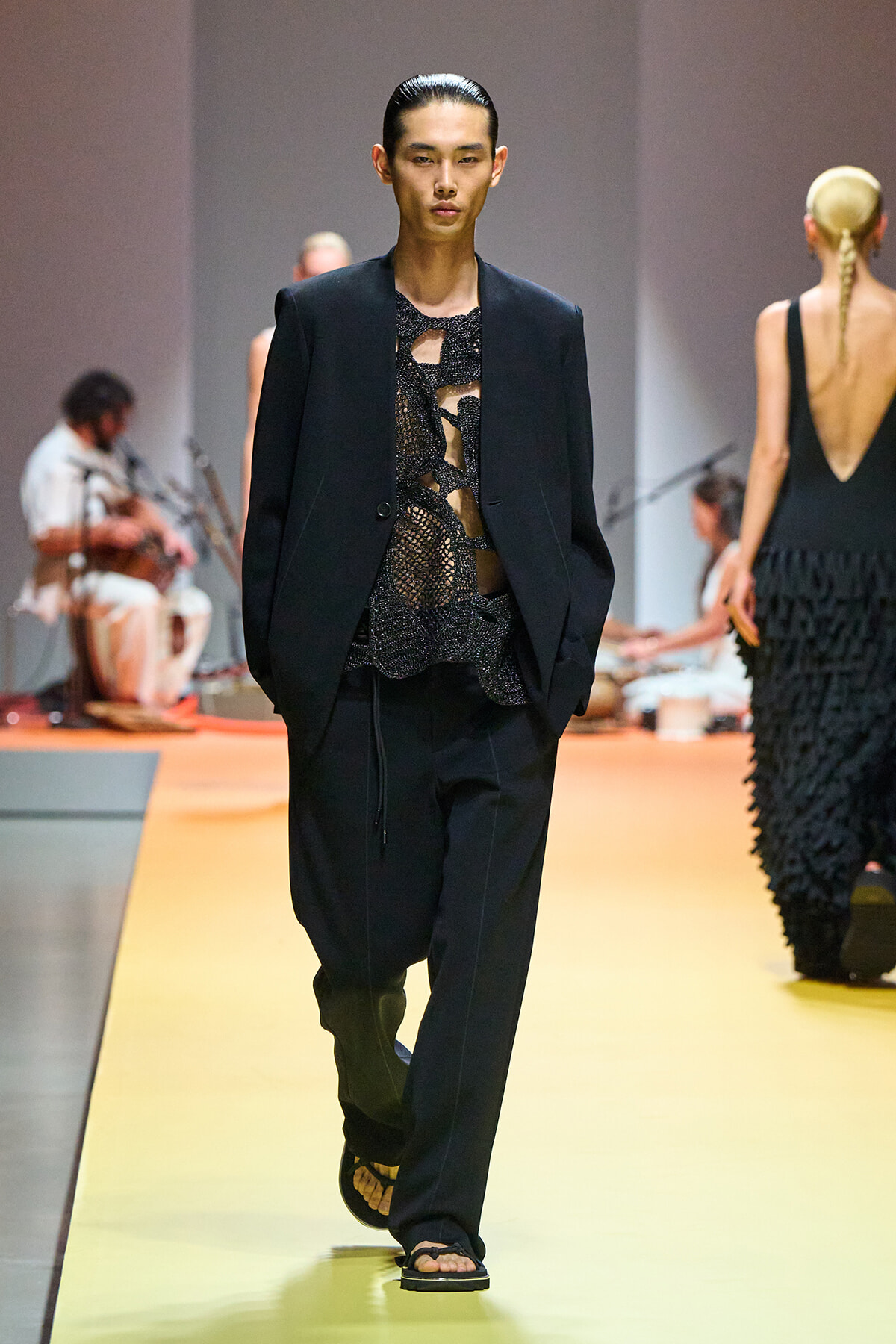 Male model walks the runway in a black blazer over a sheer, lacy top with wide black pants and black sandals on a yellow catwalk with blurred background