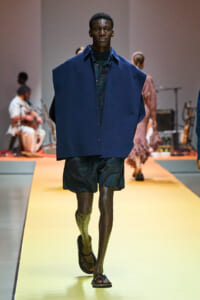 Male model walks the runway in a loose navy blue poncho over a dark shirt and dark shorts, wearing sandals.