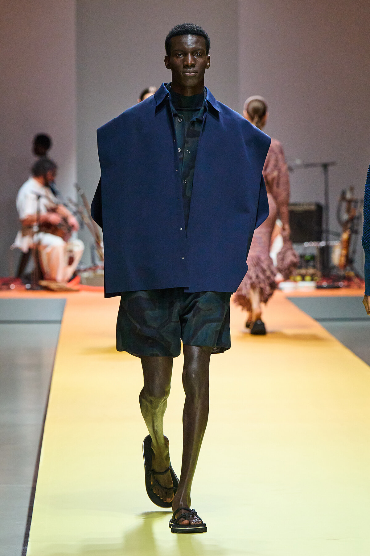 Male model walks the runway in a loose navy blue poncho over a dark shirt and dark shorts, wearing sandals.