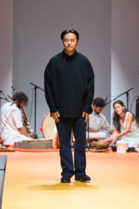 Male model in a dark, oversized shirt and navy pants standing on a yellow runway with musicians in the background southernly, performing on stage.
