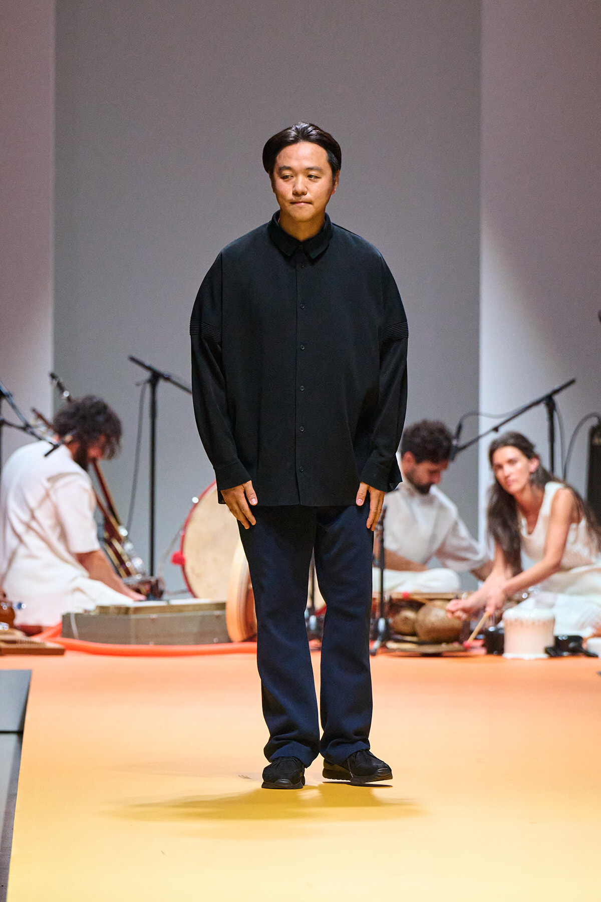 Male model in a dark, oversized shirt and navy pants standing on a yellow runway with musicians in the background southernly, performing on stage.