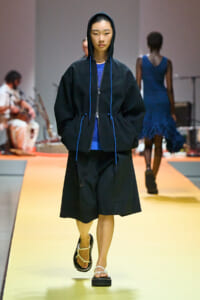 Model walks the runway in a black hooded jacket with blue drawstrings over a bright blue knitted top and knee‑length black shorts, white sandals.