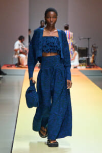 Model walking a fashion runway in a dark blue textured ensemble with a ruffled cropped top and wide-leg pants, carrying a matching bag.