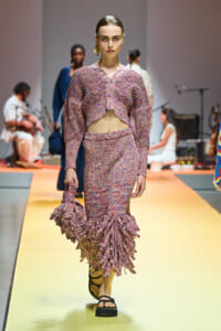 Model walks a fashion runway in a pink multicolored knit two-piece set with a cropped cardigan and ruffled hem skirt, sandals, carrying a matching ruffled bag edge.