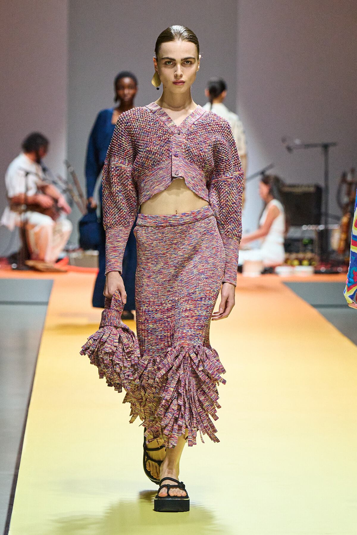 Model walks a fashion runway in a pink multicolored knit two-piece set with a cropped cardigan and ruffled hem skirt, sandals, carrying a matching ruffled bag edge.