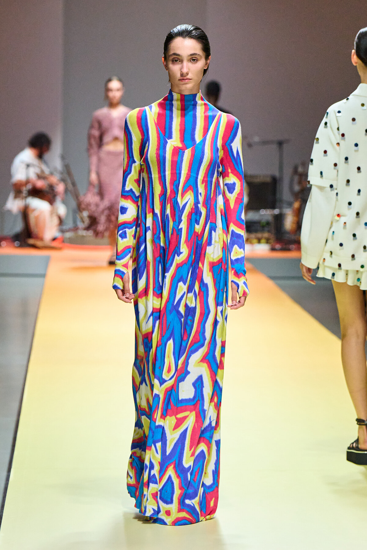 Model on a runway wearing a floor-length, high-neck dress with a bold abstract multicolor swirl pattern in blue, pink, and yellow.
