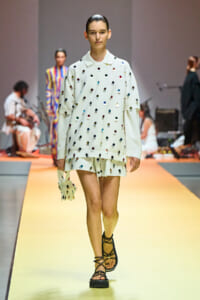Model walks the runway in a white oversized shirt and matching shorts decorated with multicolored dots, carrying a small coordinating bag with strappy sandals.]','Model on the catwalk wearing a white polka-dot outfit (top and shorts) with a tiny bag and black sandals.